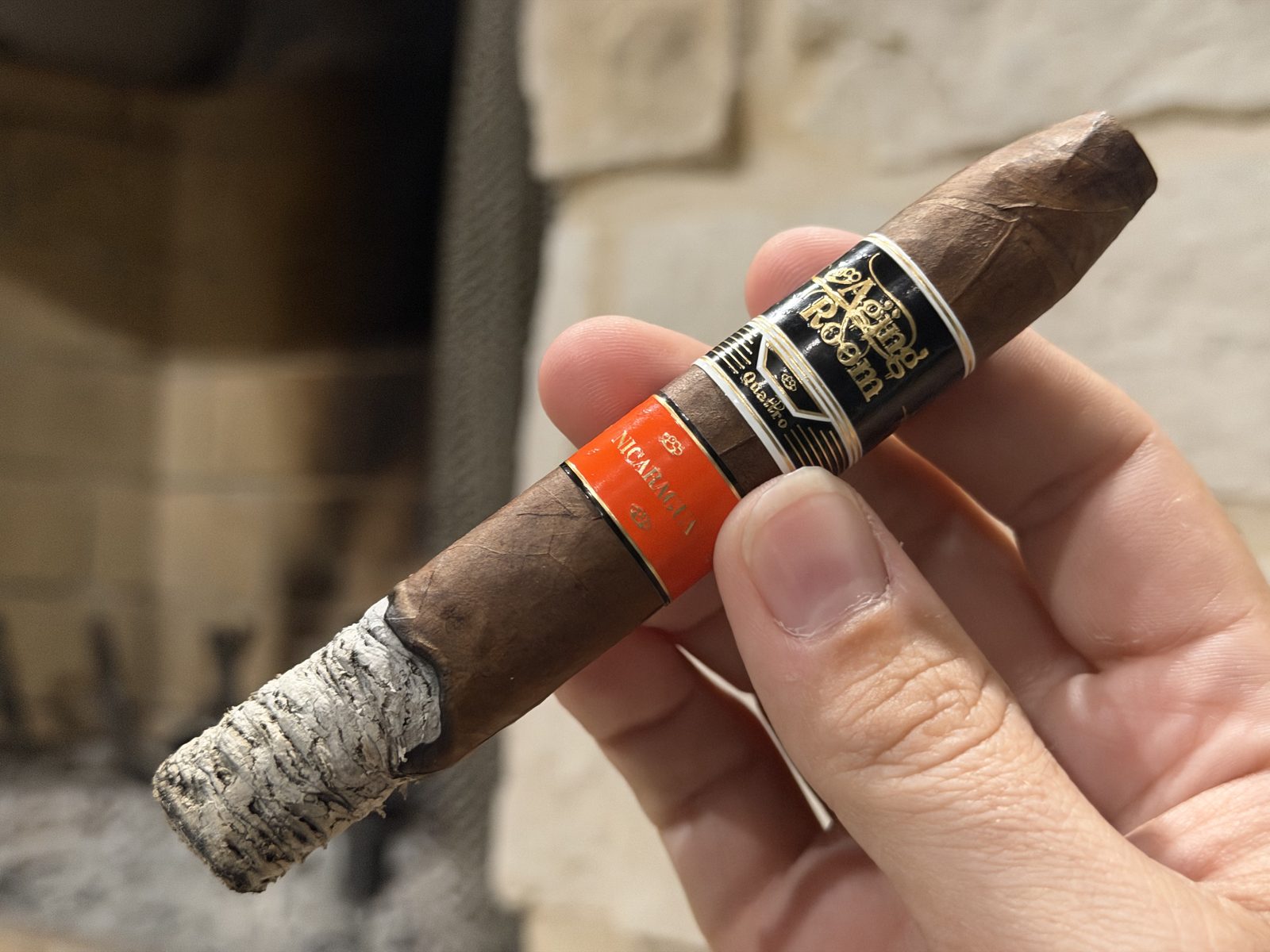 Aging Room Quattro Nicaragua Maestro at the end of the first third with about an inch of stacked gray ash, both bands still on, held in hand in front of a stone fireplace