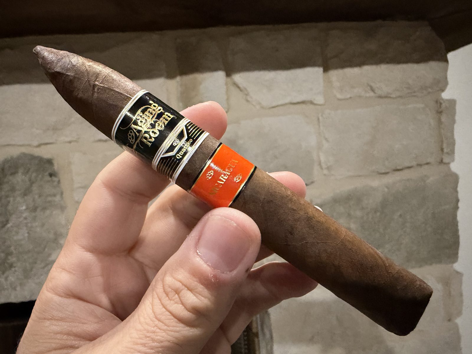 Unlit Aging Room Quattro Nicaragua Maestro box-pressed torpedo held in hand against a stone wall, dark chocolate brown wrapper with the black and gold Aging Room band and the orange Nicaragua secondary band