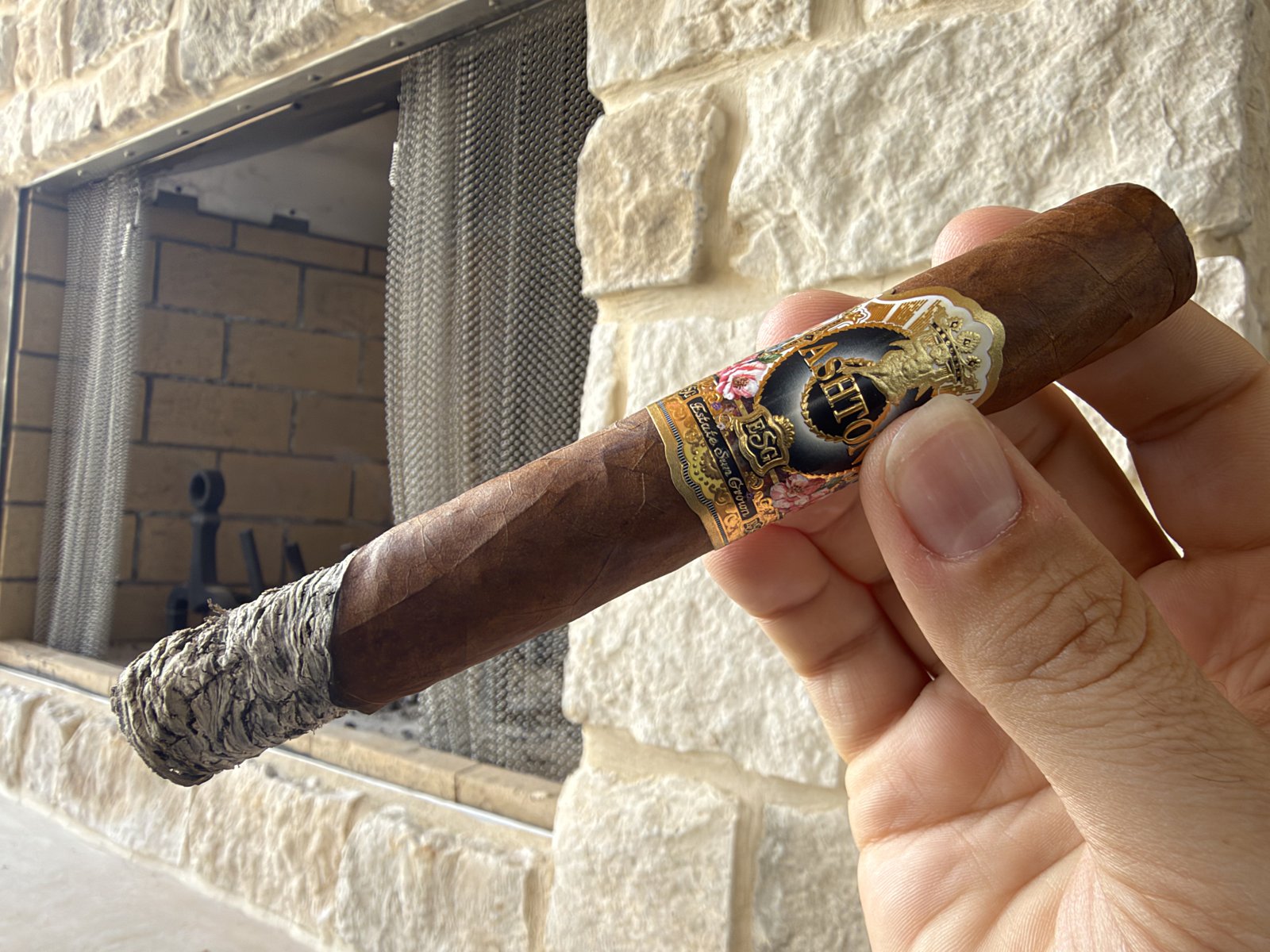 Ashton ESG 23 Year Salute Toro burning near the end of the first third, a long gray ash stack with a visibly crooked burn line, held in hand in front of a stone fireplace