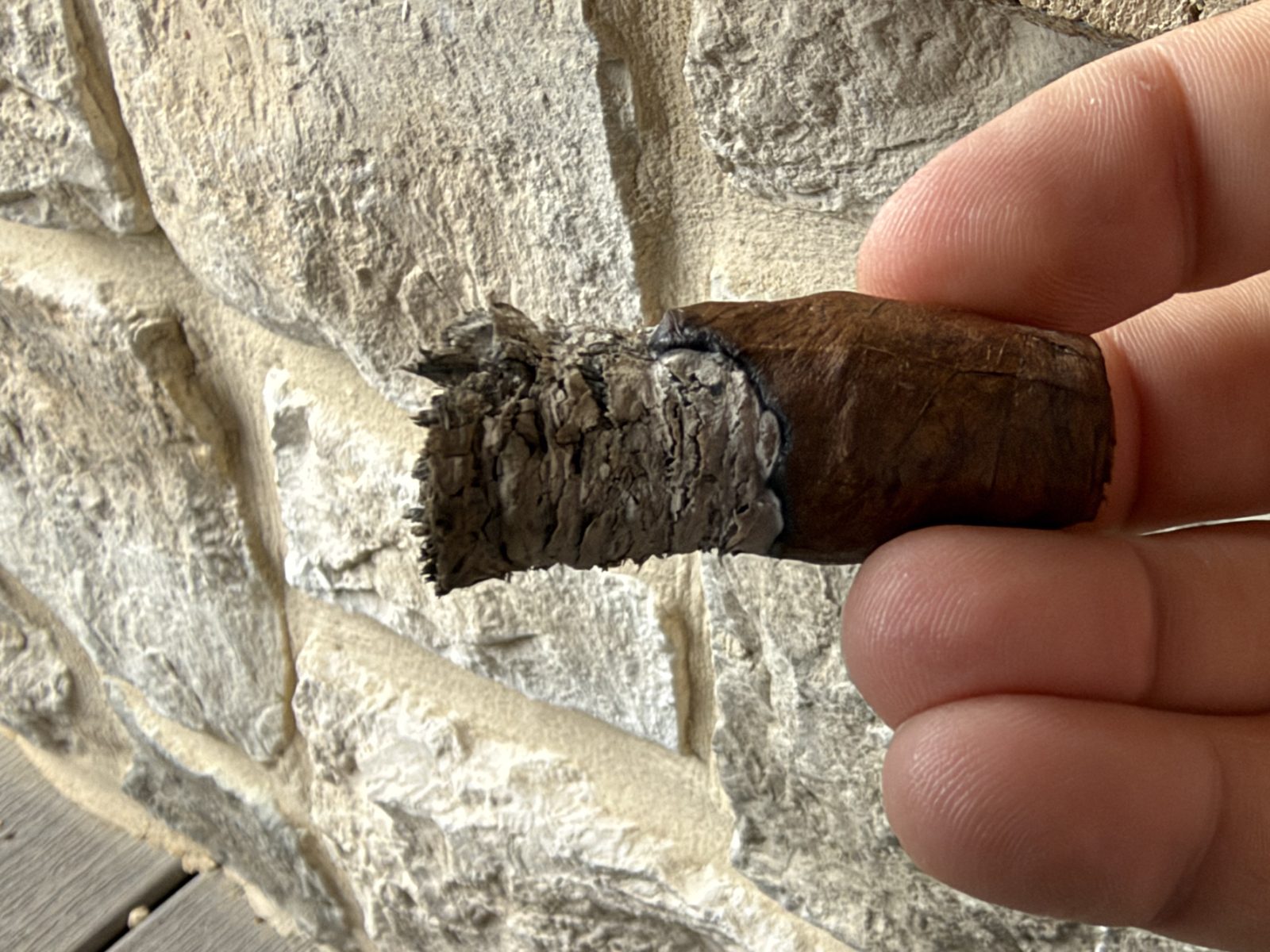 Nub of the Ashton VSG Torpedo held between fingers at the end of the smoke, a short ragged ash and a dark wrapper with no band, in front of a stone wall