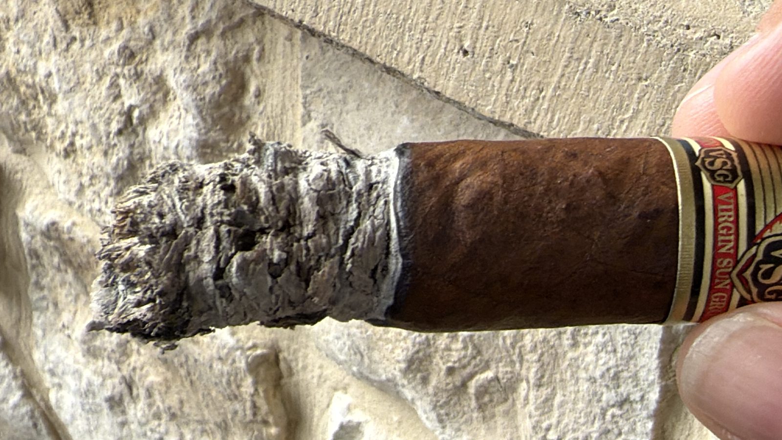 Ashton VSG Torpedo at the end of the second third, a ragged gray ash cracking along one side, band still on and reading Virgin Sun Grown, held in fingers in front of a stone wall