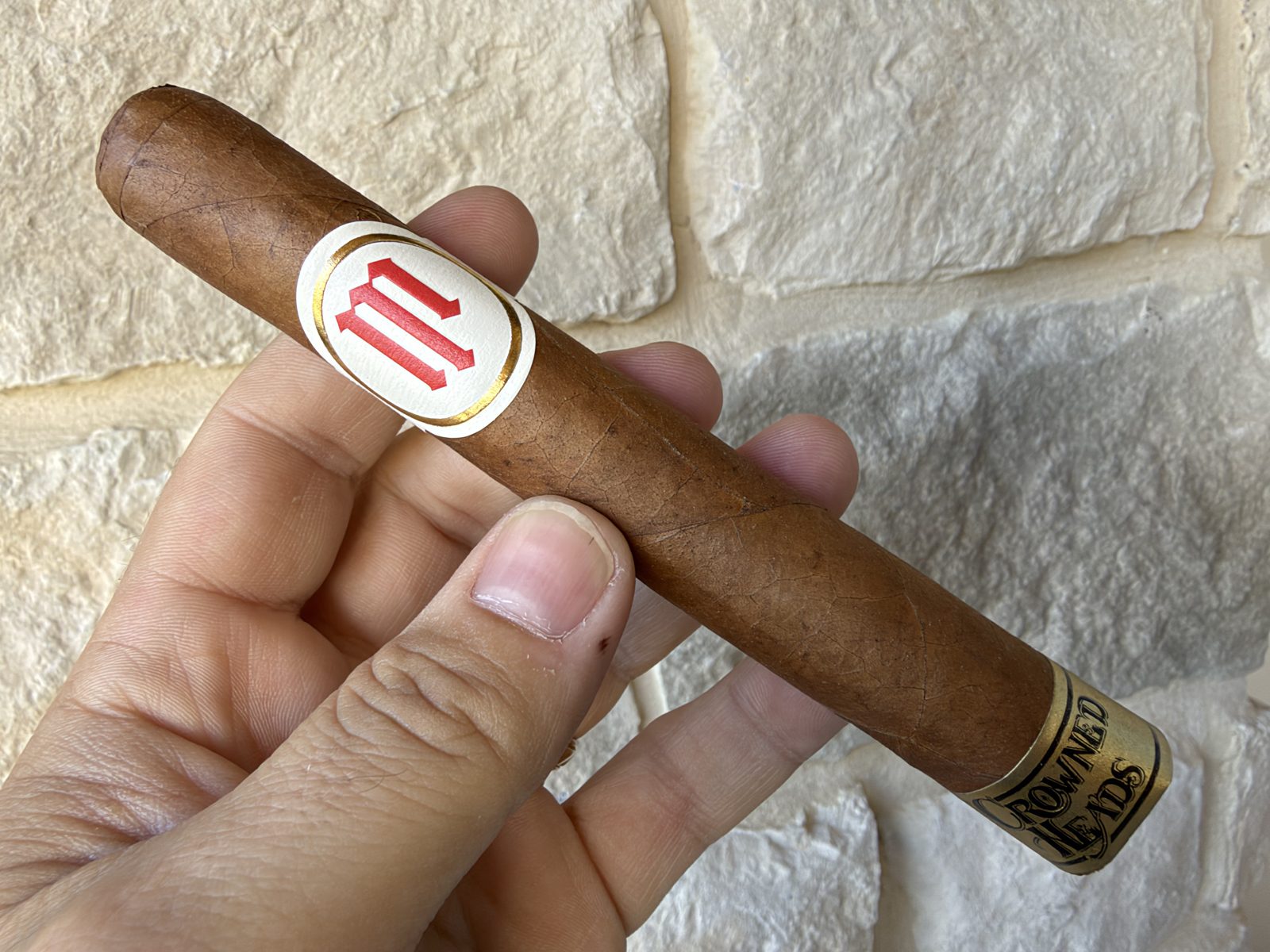 Unlit Crowned Heads Mil Días Sublime held in hand against a stone wall, light brown Ecuadorian Habano wrapper with the white and red Mil Días band and the gold Crowned Heads foot band