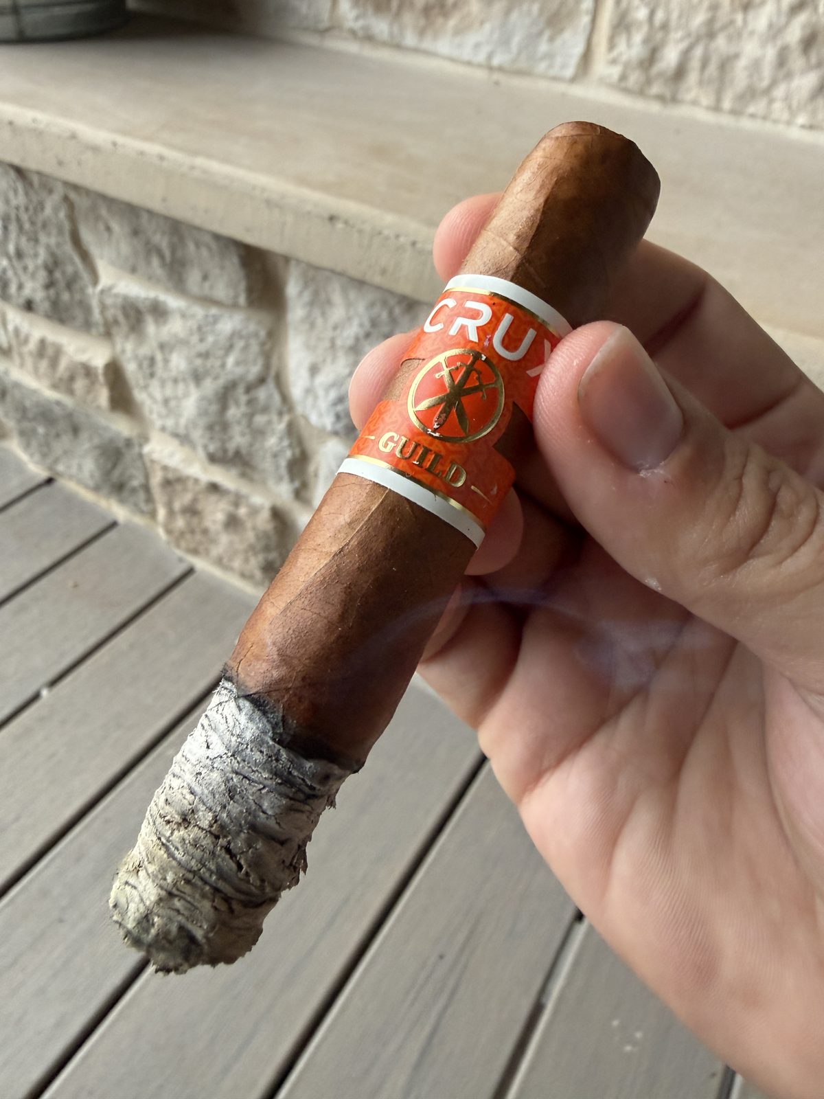 Crux Guild Toro held in hand, orange and gold Guild band, stacked gray ash at the foot and a wisp of smoke rising