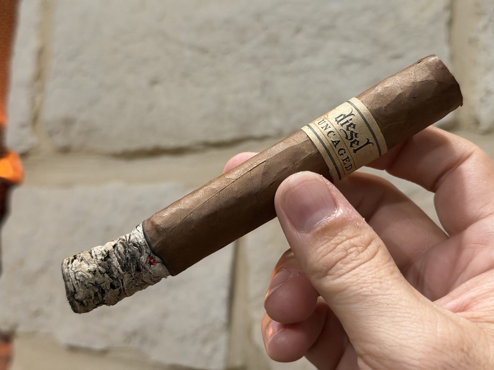 Diesel Uncaged Toro at the end of the first third, holding a long, layered gray ash, band still on, in front of a stone wall