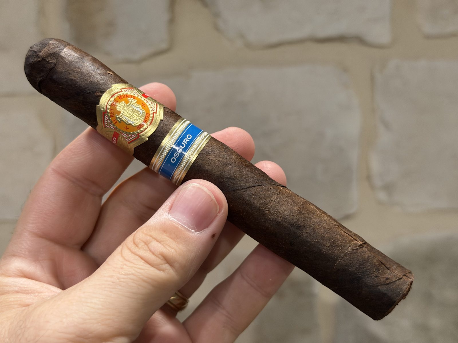 Unlit El Rey del Mundo Oscuro Robusto Larga out of the tissue, held in hand against a stone wall, dark brown Connecticut Broadleaf Oscuro wrapper with thick veins and the two bands