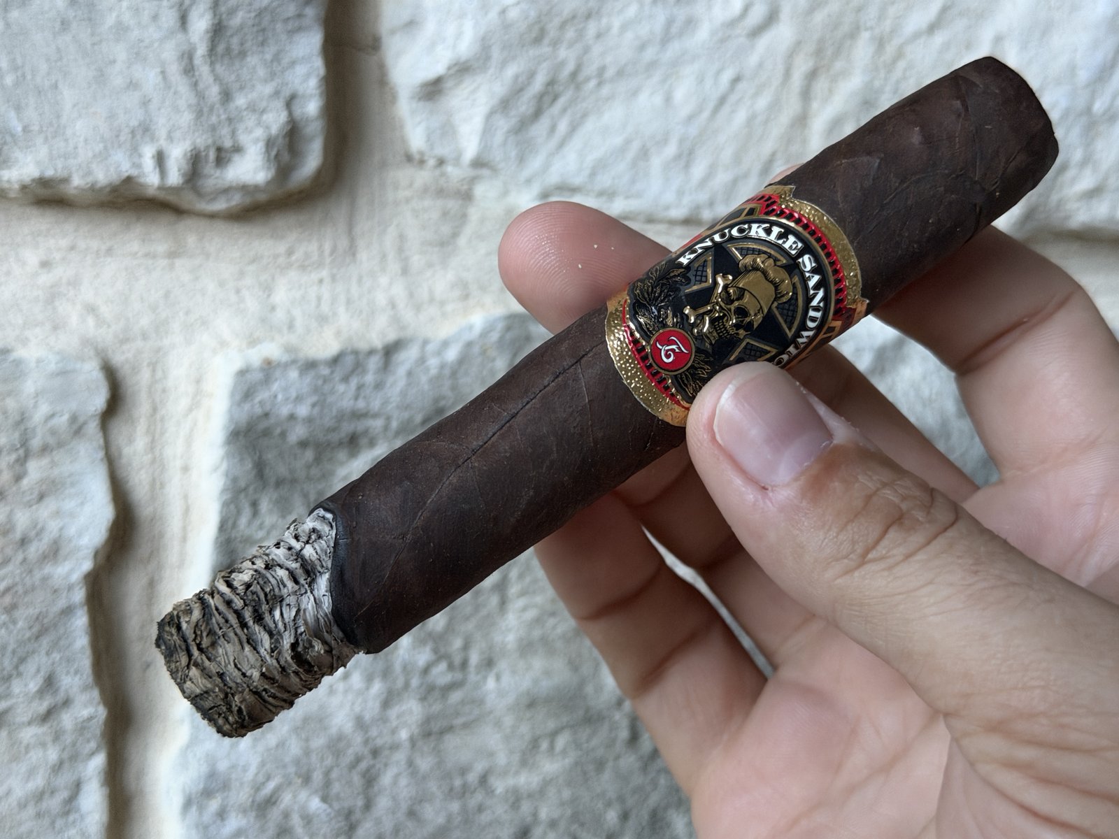 Lit Espinosa Knuckle Sandwich with a long, tight, light-grey stacked ash at the foot, band still on, held against a stone wall