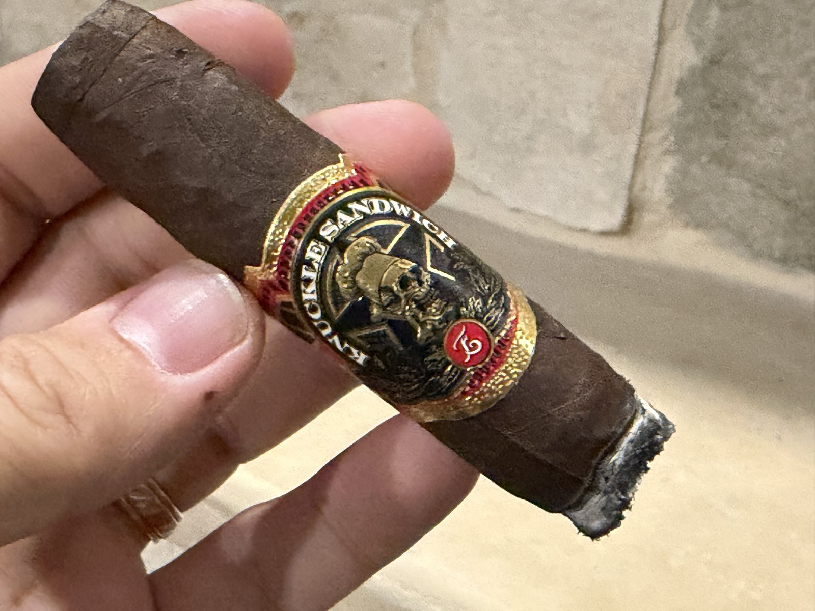 Lit Espinosa Knuckle Sandwich with the black-and-gold skull band readable, short ash and a glowing ember at the foot, held against a stone wall