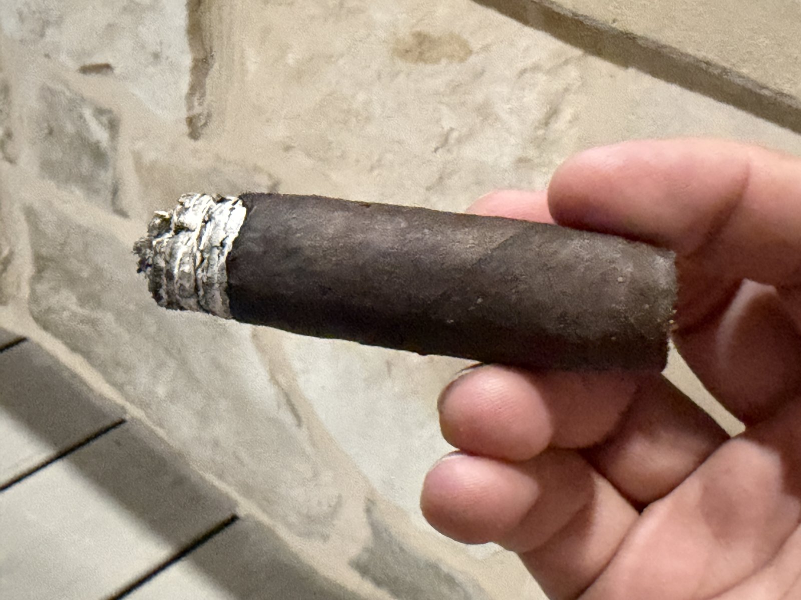 Espinosa Knuckle Sandwich burned down into the final third with the band removed, dark wrapper and a lit ember at the foot, held against a stone wall