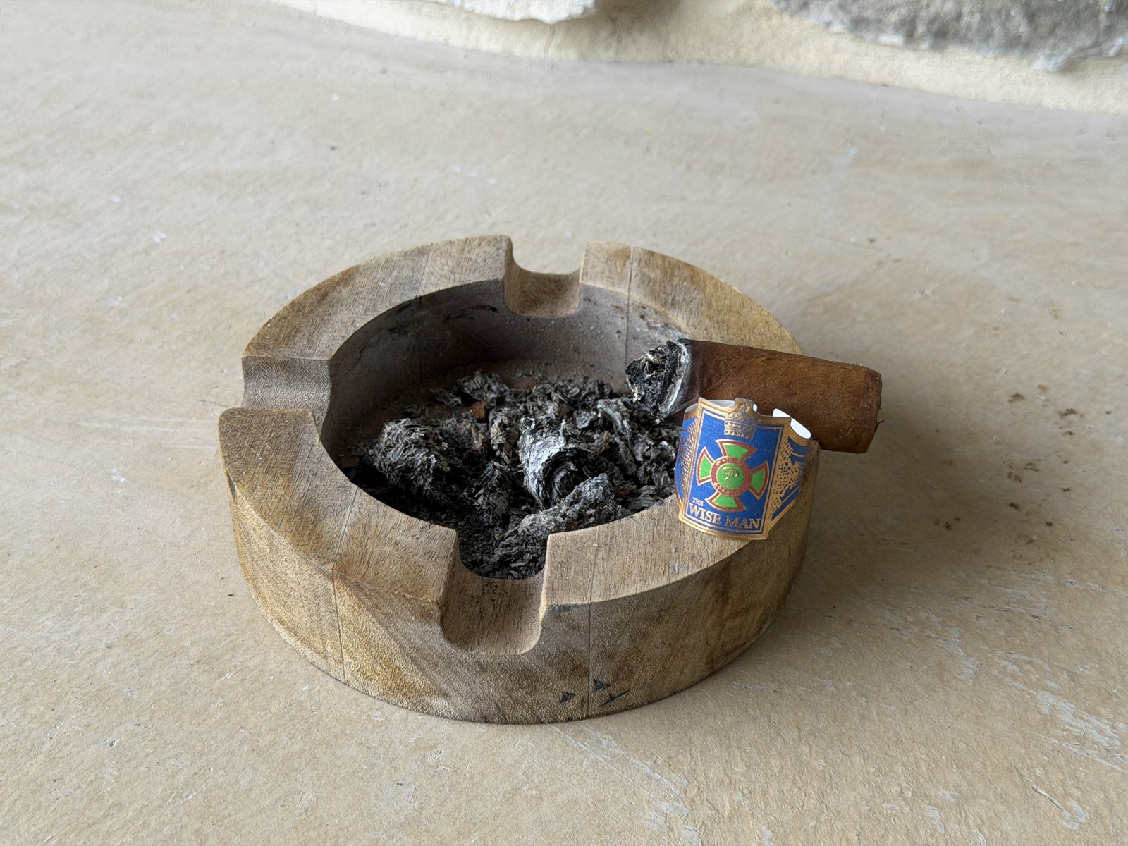 The finished Wise Man Corojo nub resting on a wooden ashtray full of loose flaky ash, band sitting beside it