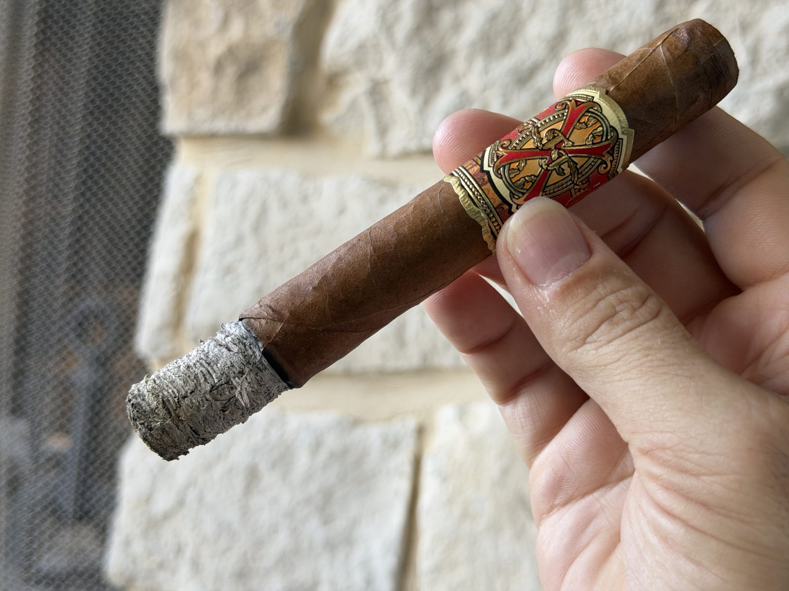 Fuente Fuente Opus X Perfecxion X burning with a light gray ash stack at the end of the first third, band still on, held in hand in front of a stone wall
