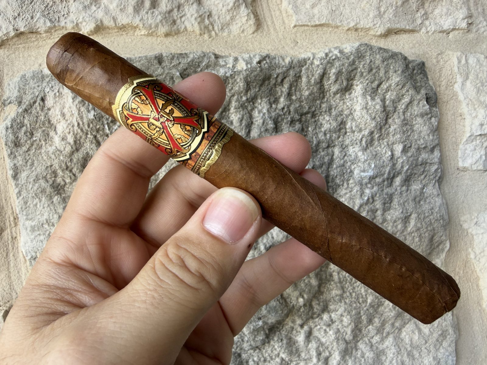 Unlit Fuente Fuente Opus X Perfecxion X held in hand against a stone wall, medium brown rosado wrapper with the red and gold Opus X band