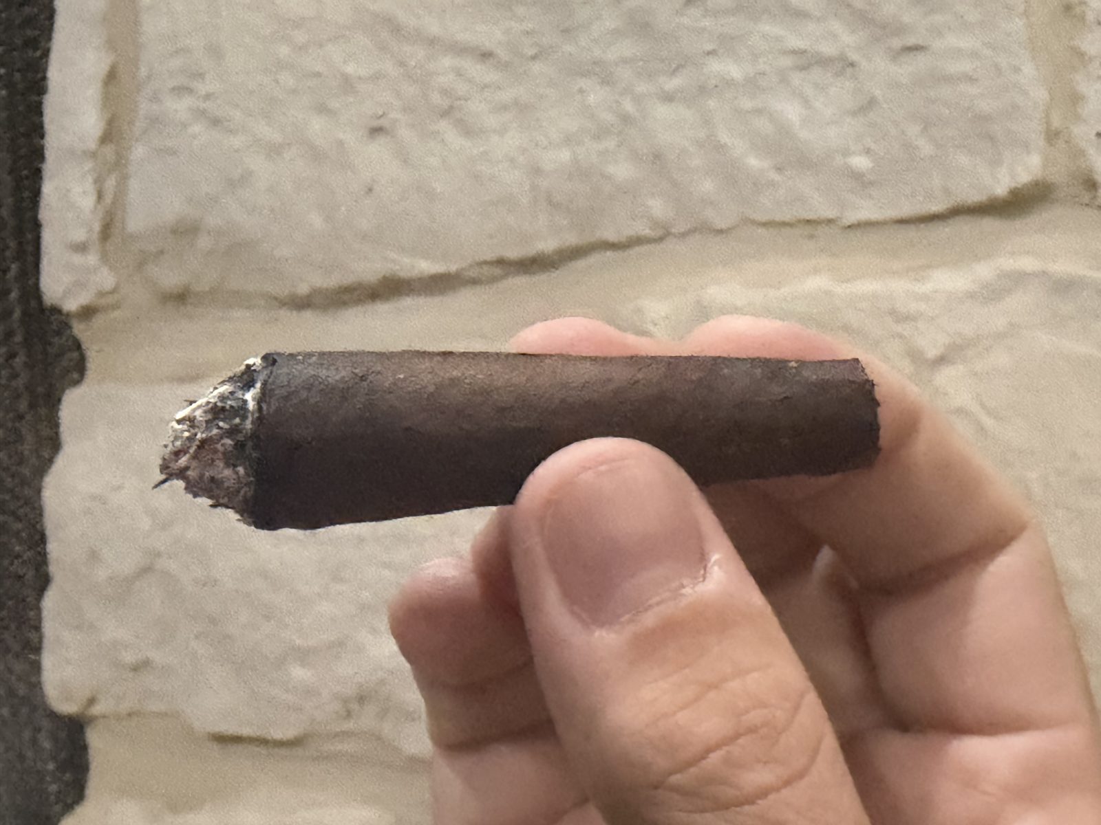 Liga Privada Unico Serie Nasty Fritas with the ash knocked off, short burned foot and the narrow tapered body held in fingertips against a stone wall