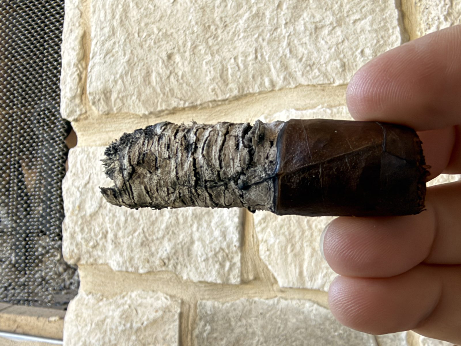 Liga Undercrown Maduro Gran Toro late in the final third with a long, tight, layered gray ash and a nearly straight burn line, held in fingertips in front of a stone outdoor fireplace