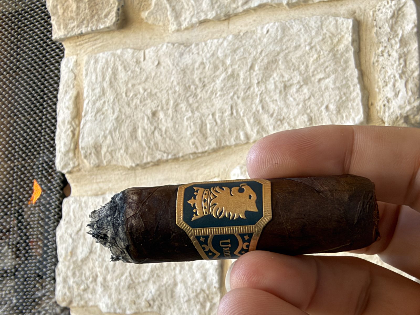 Liga Undercrown Maduro Gran Toro just after ashing at the end of the second third, showing a raised ligero point in the center of the foot, gold and teal band on the cigar, in front of a stone outdoor fireplace
