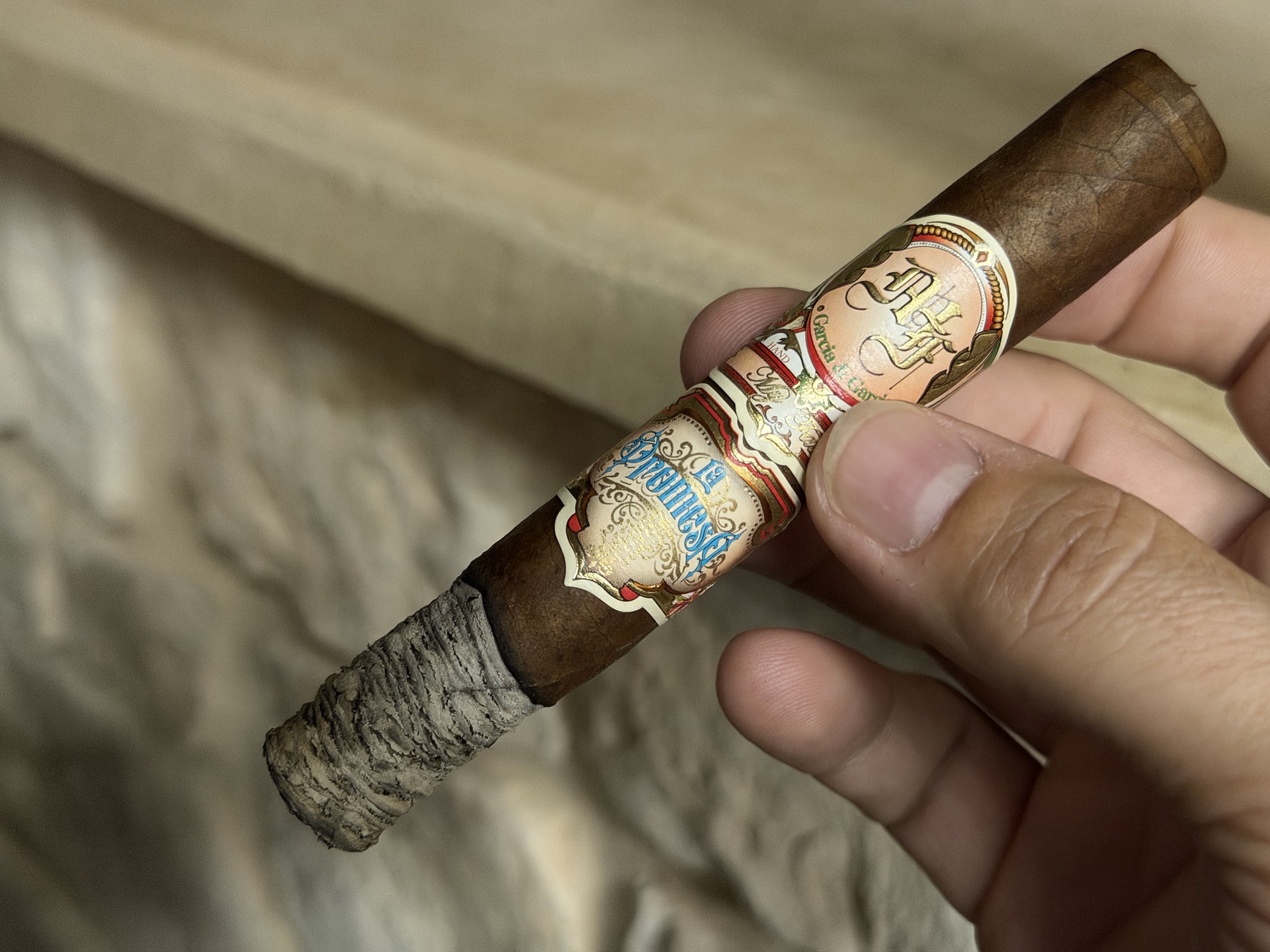 La Promesa burning in the first third with a tight gray stacked ash, both bands still on, against a stone wall