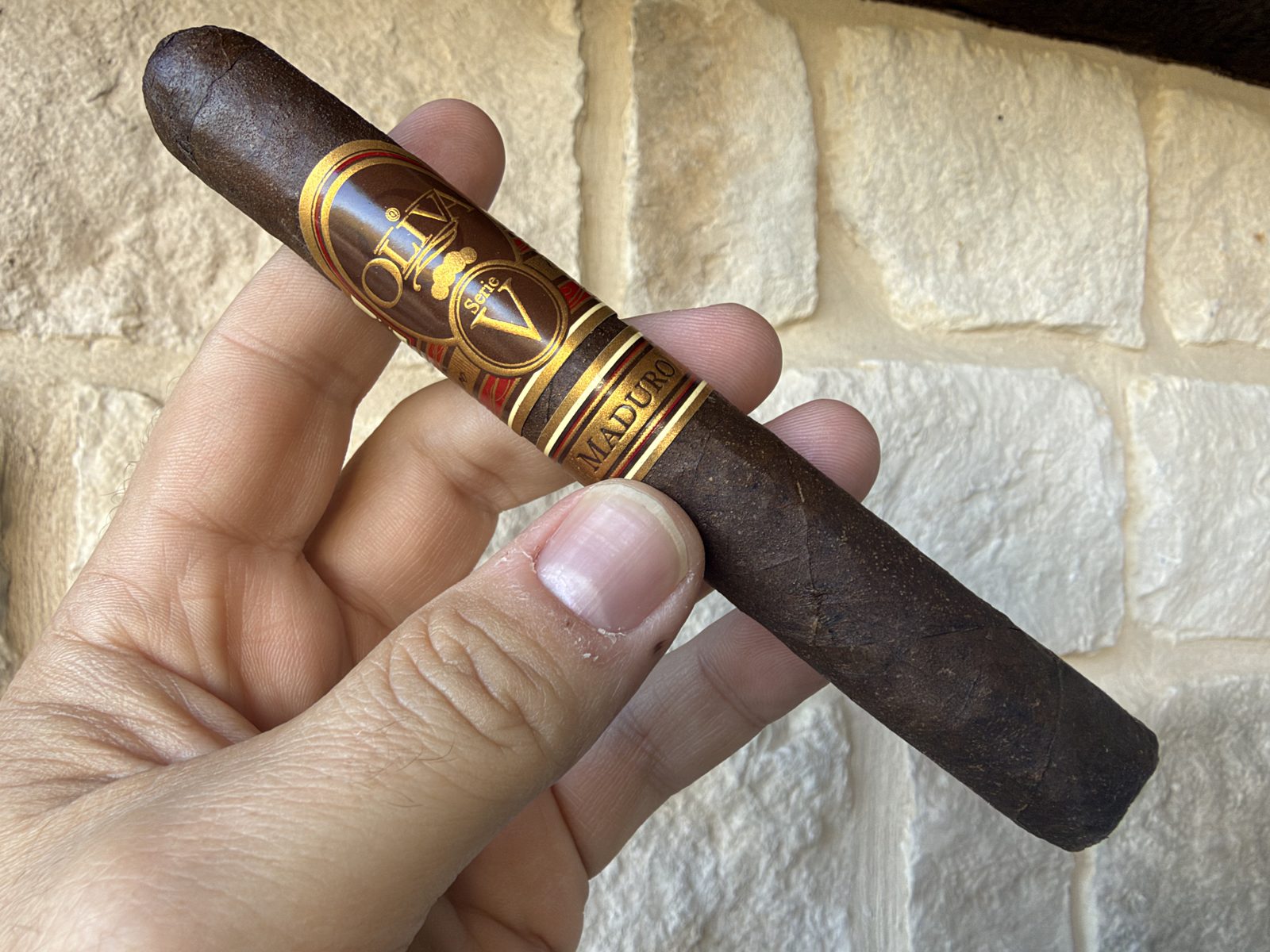 Unlit Oliva Serie V Maduro Toro held in hand against a stone wall, very dark brown lightly toothy wrapper with the red and gold Serie V band and the secondary Maduro band