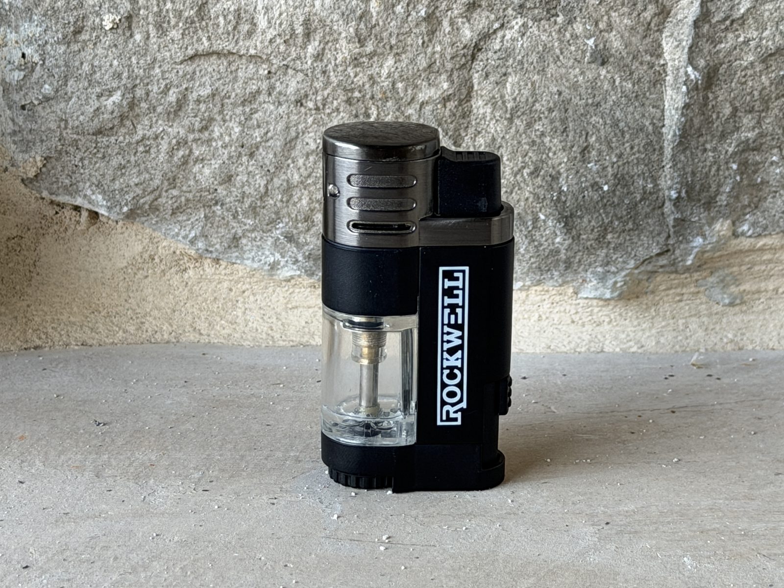 Rockwell quad torch pocket lighter standing upright, black plastic body with clear fuel tank window and Rockwell branding