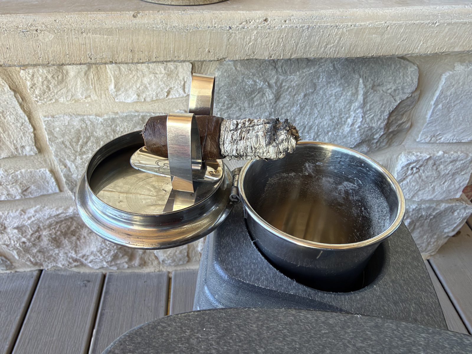 Polished stainless steel Stinky Cigar Car Ashtray sitting in a dark gray outdoor cupholder, lid open to the side with a lit Liga Undercrown Maduro held in the two stainless cigar wings