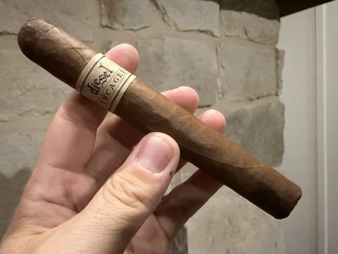Unlit Diesel Uncaged Toro held in hand against a stone wall, light brown Habano Criollo wrapper with the cream paper Diesel Uncaged band