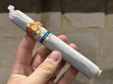 El Rey del Mundo Oscuro Robusto Larga still wrapped in its white tissue paper, held in hand against a stone wall, with the orange and gold El Rey del Mundo band and the blue Oscuro secondary band showing through