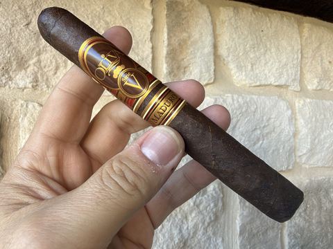 Unlit Oliva Serie V Maduro Toro held in hand against a stone wall, very dark brown lightly toothy wrapper with the red and gold Serie V band and the secondary Maduro band