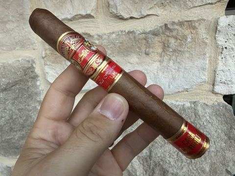 Unlit Perez-Carrillo La Historia Silk Toro held in hand against a stone wall, light brown wrapper with red and gold main band, secondary La Historia Silk band, and Casa Carrillo foot band
