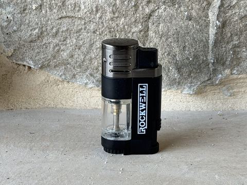 Rockwell quad torch pocket lighter standing upright, black plastic body with clear fuel tank window and Rockwell branding
