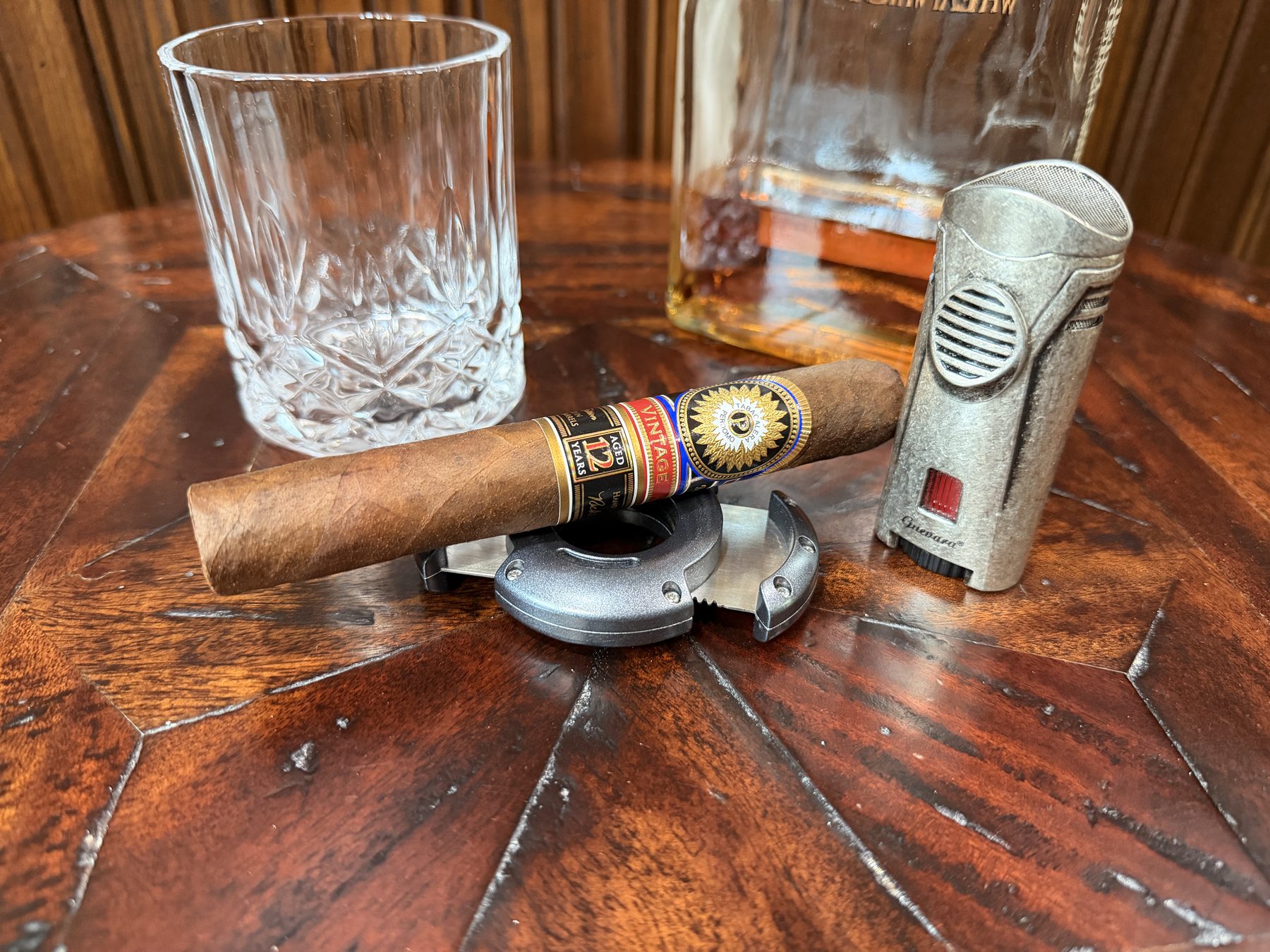 Cigar resting on a cutter beside a torch lighter and a glass of bourbon on a dark wood table