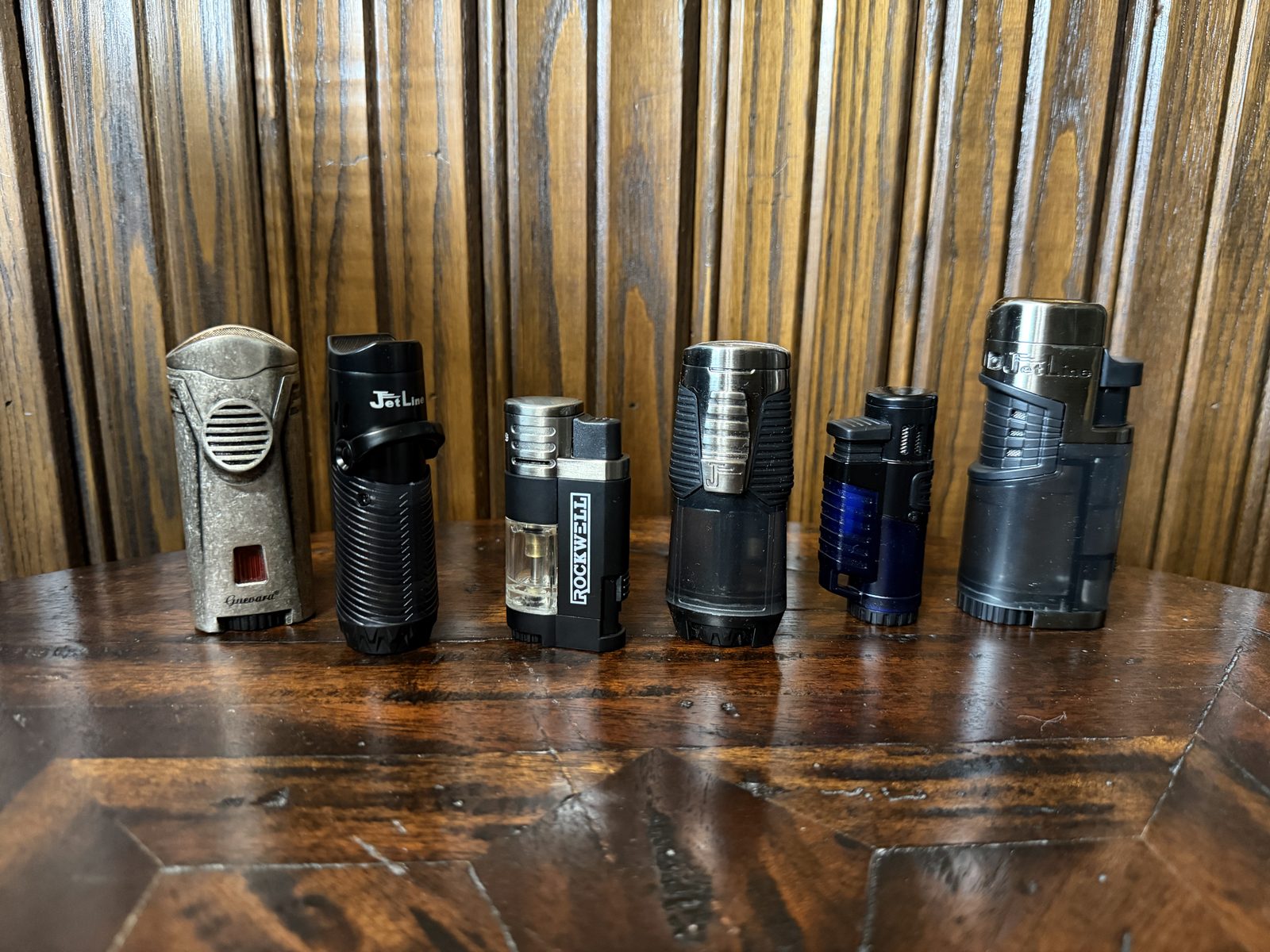 Six torch lighters lined up on a dark wood table against wood paneling